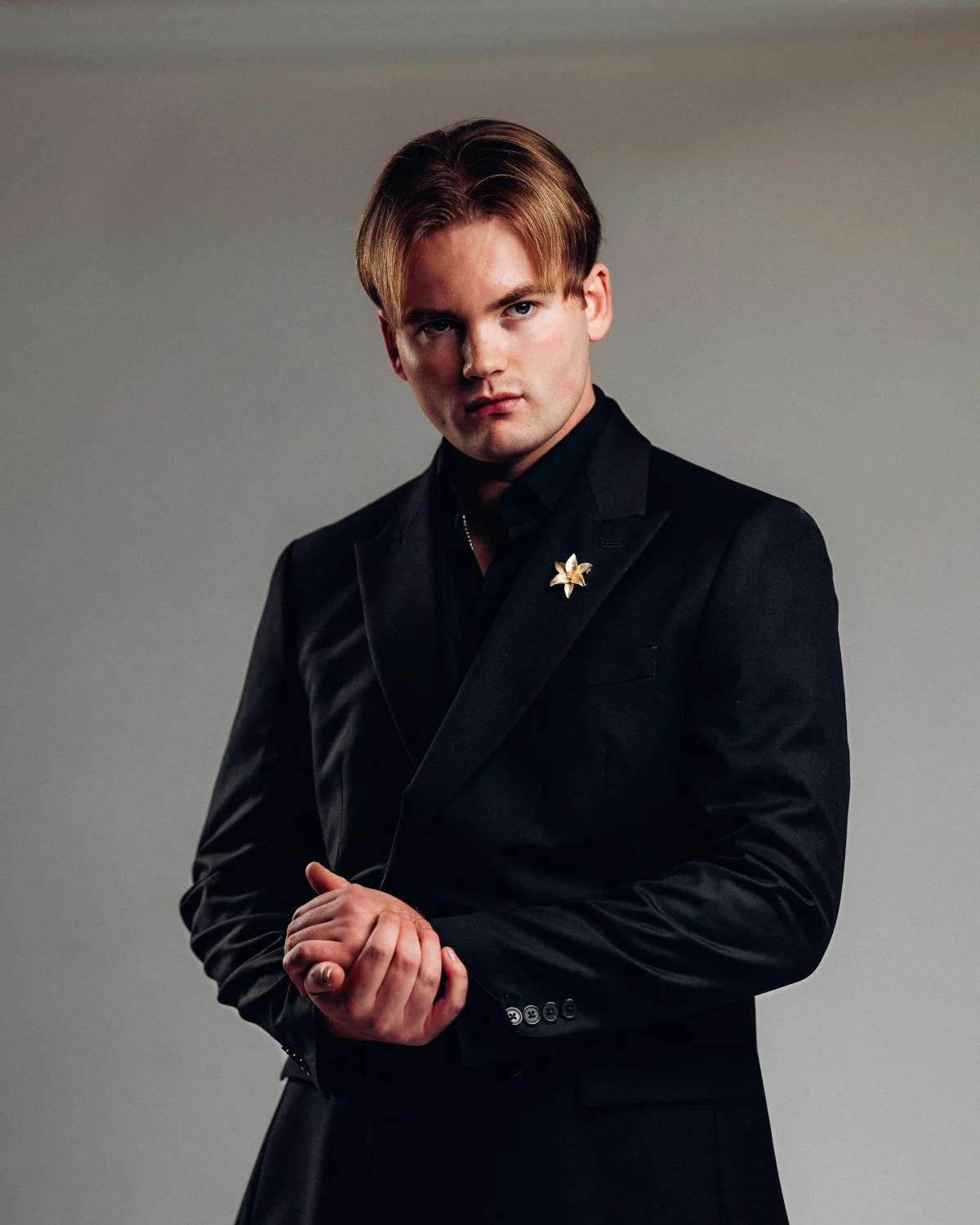 A man with blond hair wearing a black suit and gold star pin, posing against a neutral grey studio background.