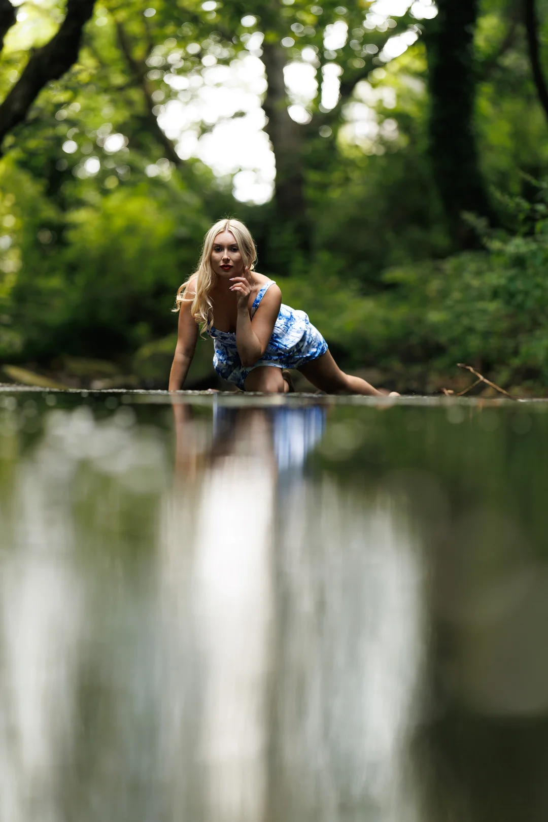Blonde woman in a blue dress posing by a reflective pond in a green forest.