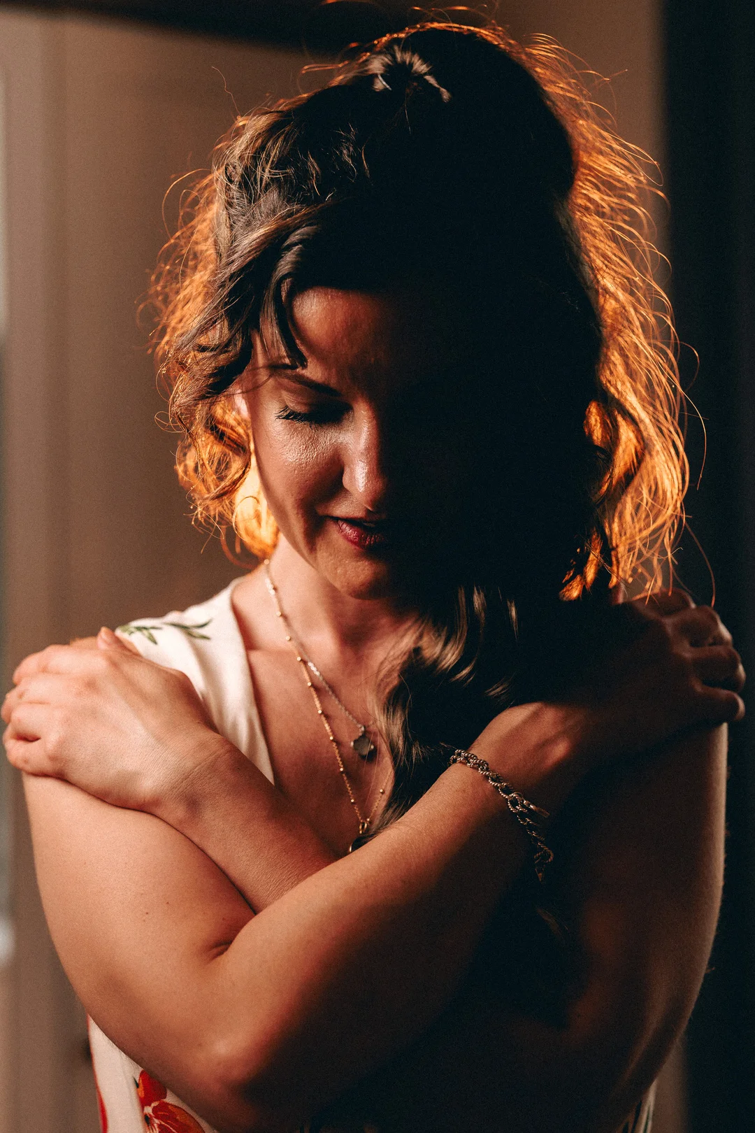 Moody portrait of a woman with curly hair hugging herself, illuminated by warm rim lighting.