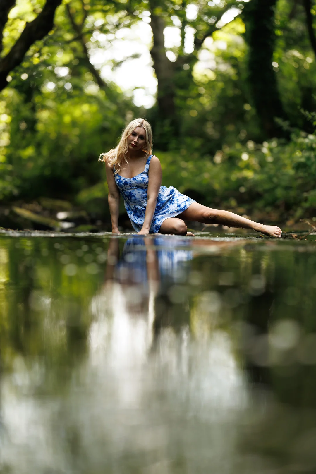 Blonde woman in a blue floral dress posing by a forest stream with a reflection in the foreground.