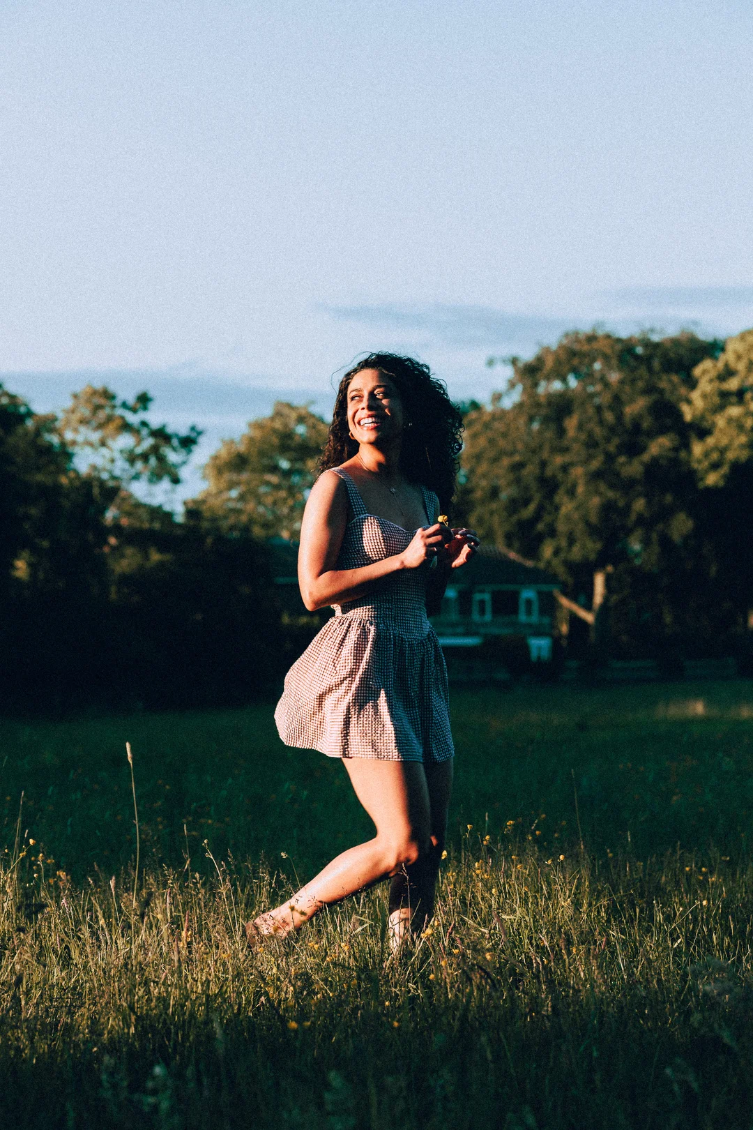 A smiling woman with curly hair in a red and white gingham dress standing in a sunlit grassy field.