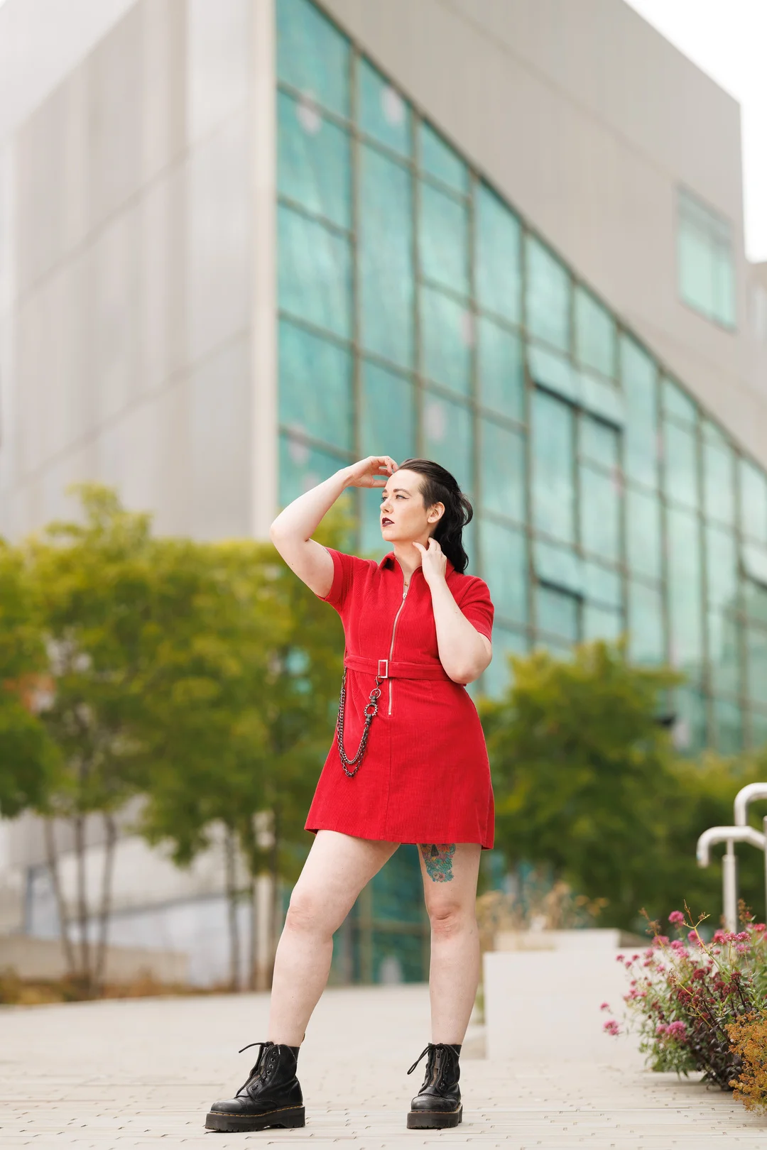 A woman in a short red dress and black boots posing in front of a modern glass building.