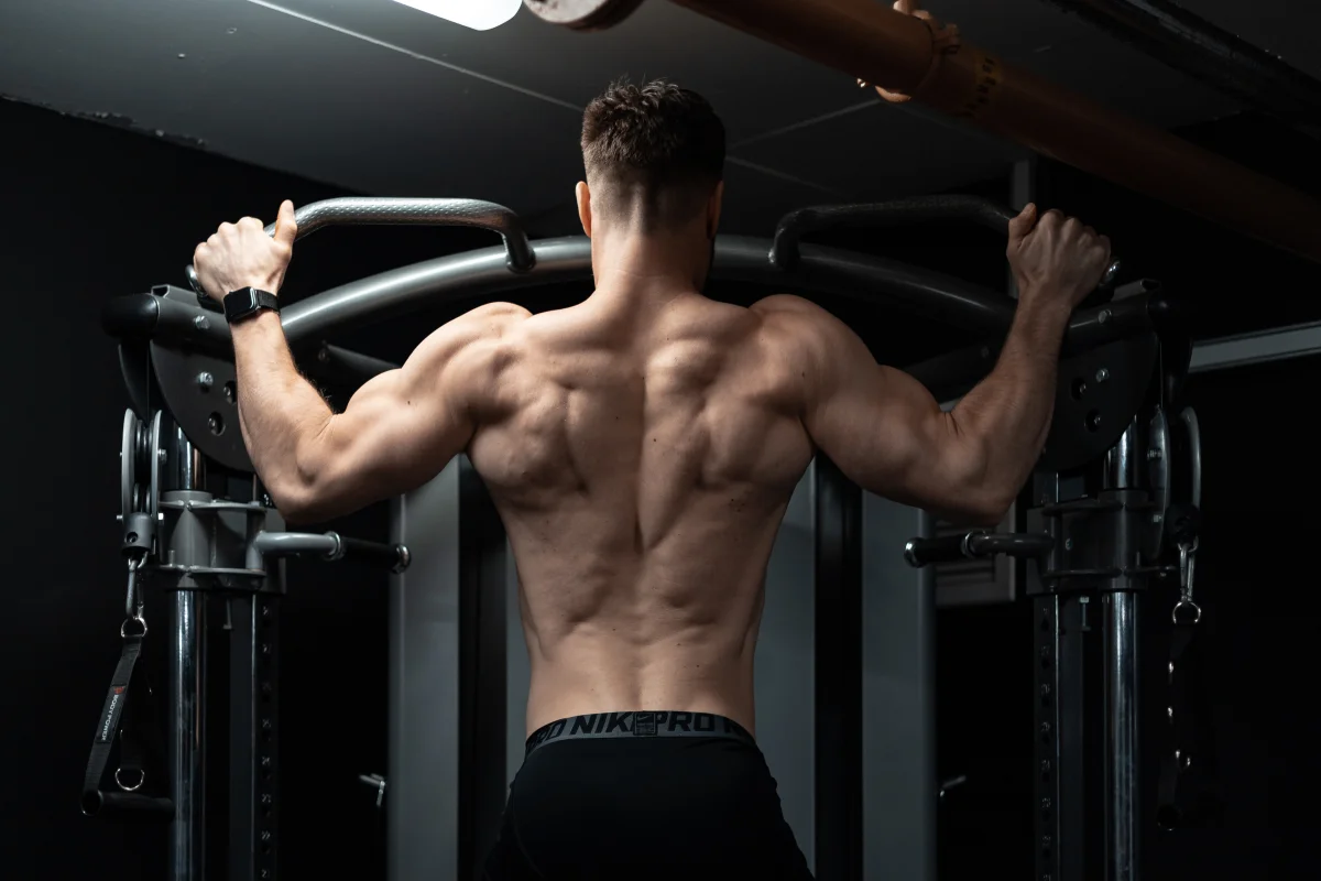 Rear view of a muscular man flexing his back muscles on a gym machine in a moody, dark setting.