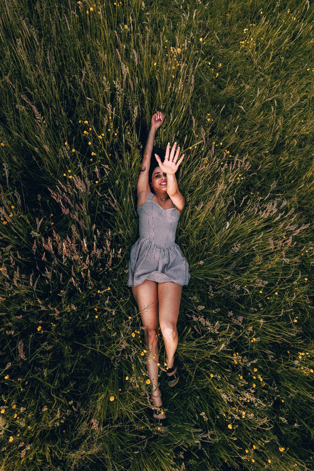 Woman in a checkered dress lying in a field of tall green grass and yellow wildflowers, smiling.