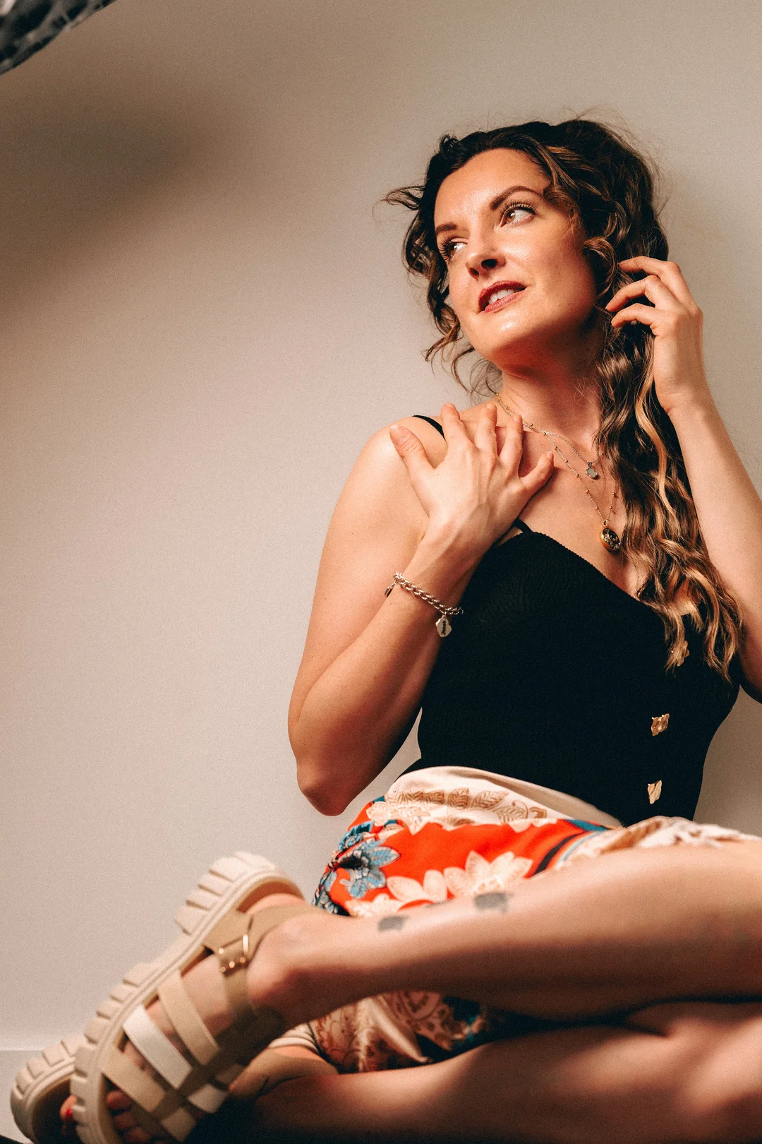 A woman with wavy hair in a black top and colorful skirt posing against a neutral wall.