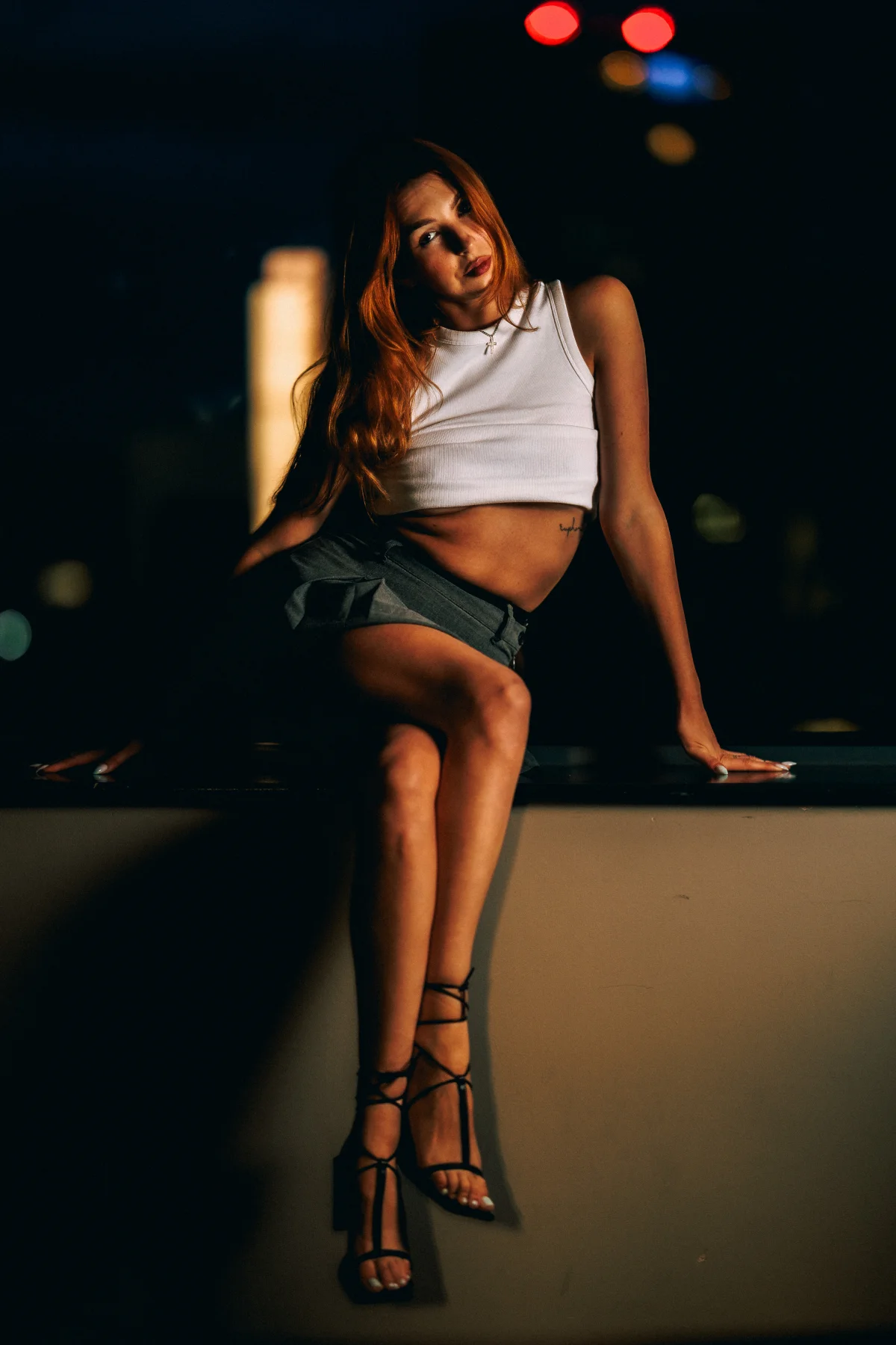 Woman with red hair posing on a ledge at night in a white crop top and grey skirt.
