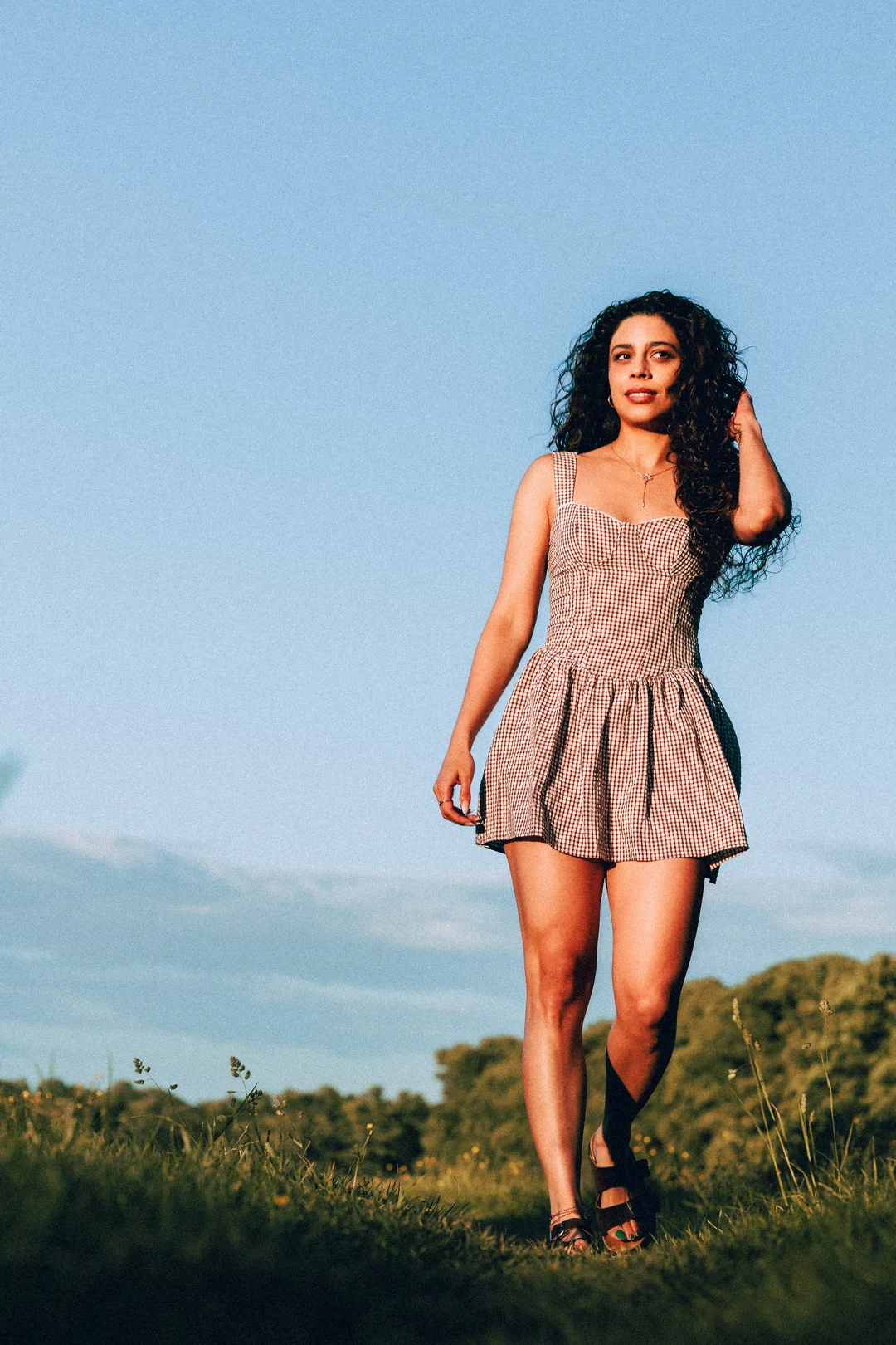 Woman with curly hair in a red and white checkered dress standing in a grassy field under a blue sky.