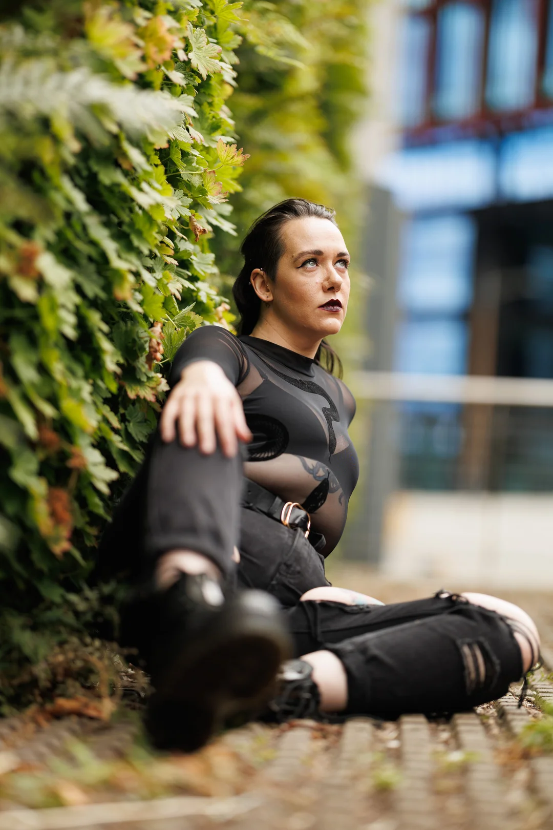 A person with dark hair and makeup sitting against a green hedge, wearing a sheer black top and ripped black pants.