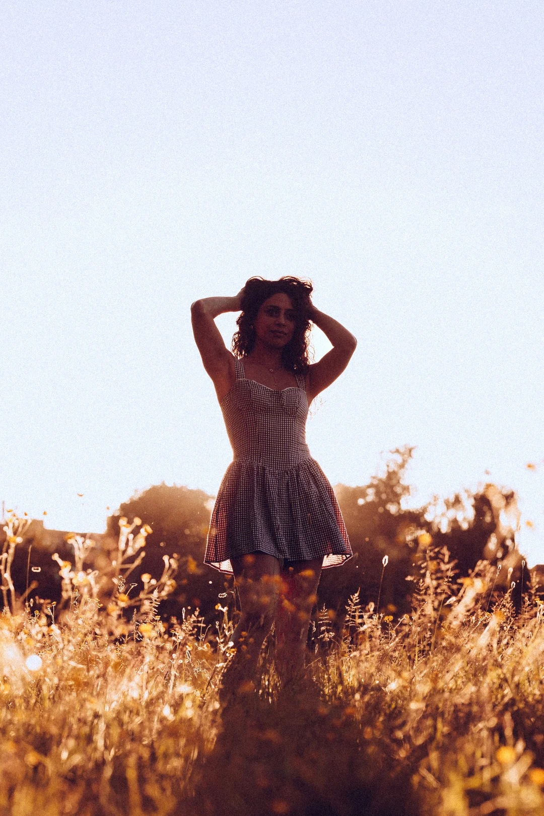 A woman in a red and white checkered dress posing in a golden field during sunset.