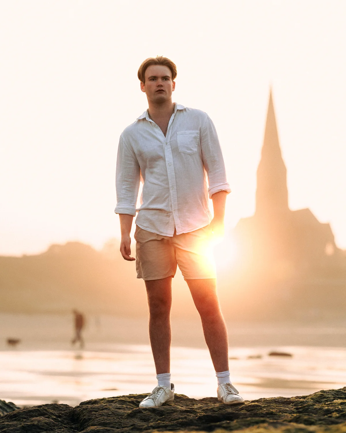 Full-body portrait of a man in white linen clothing standing on a rocky beach during a golden hour sunset.