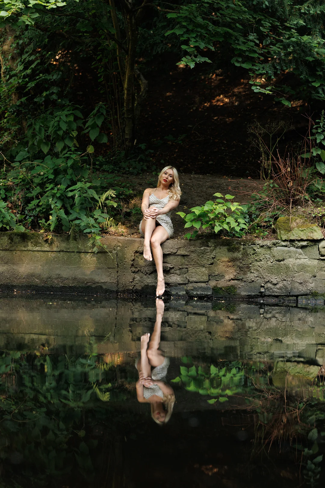 A woman in a short silver dress sitting on a stone ledge by a still pond, creating a clear reflection.