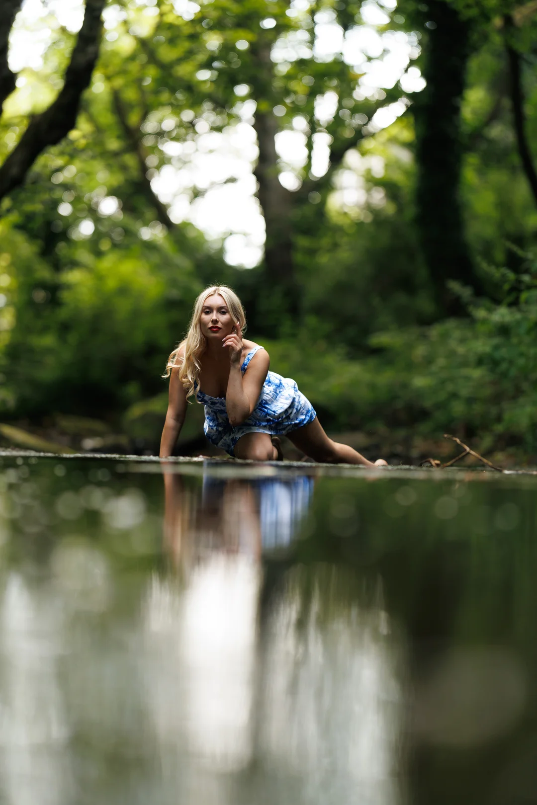 Blonde woman in a blue dress posing by a reflective pool in a lush green forest.