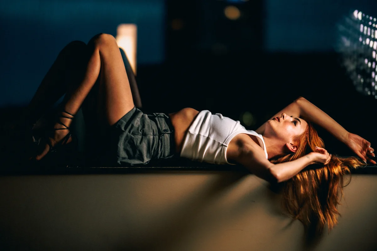 Woman with red hair lying on a ledge at night in a white crop top and grey shorts, moody urban lighting.