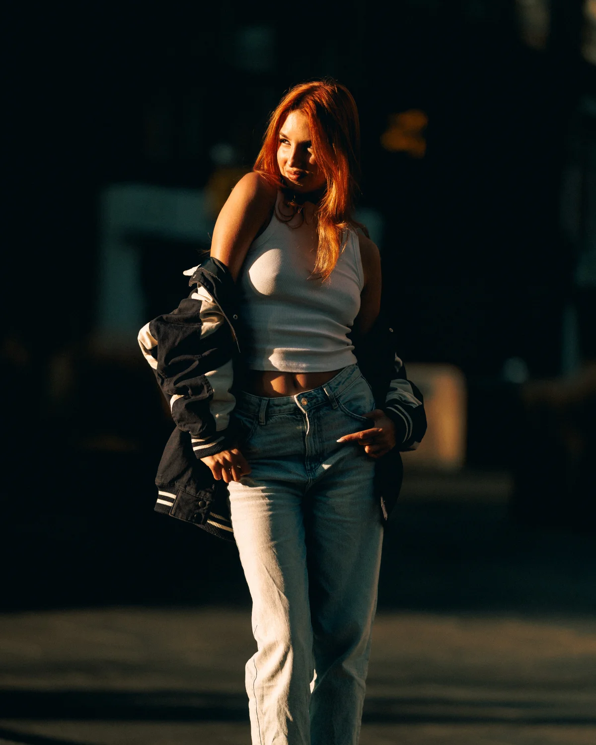 Young woman with red hair posing in a white crop top, jeans, and varsity jacket during golden hour.