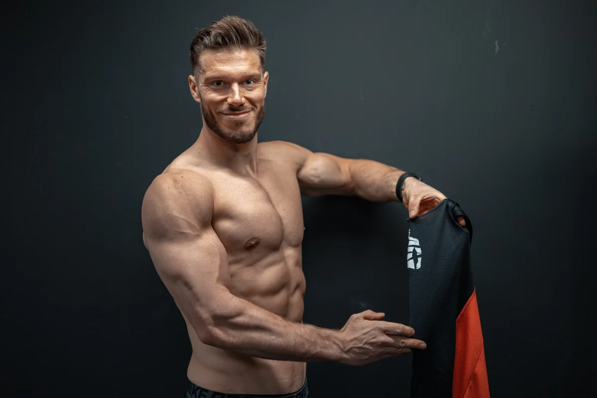 Shirtless muscular man with a beard holding a black and red athletic shirt against a dark background.