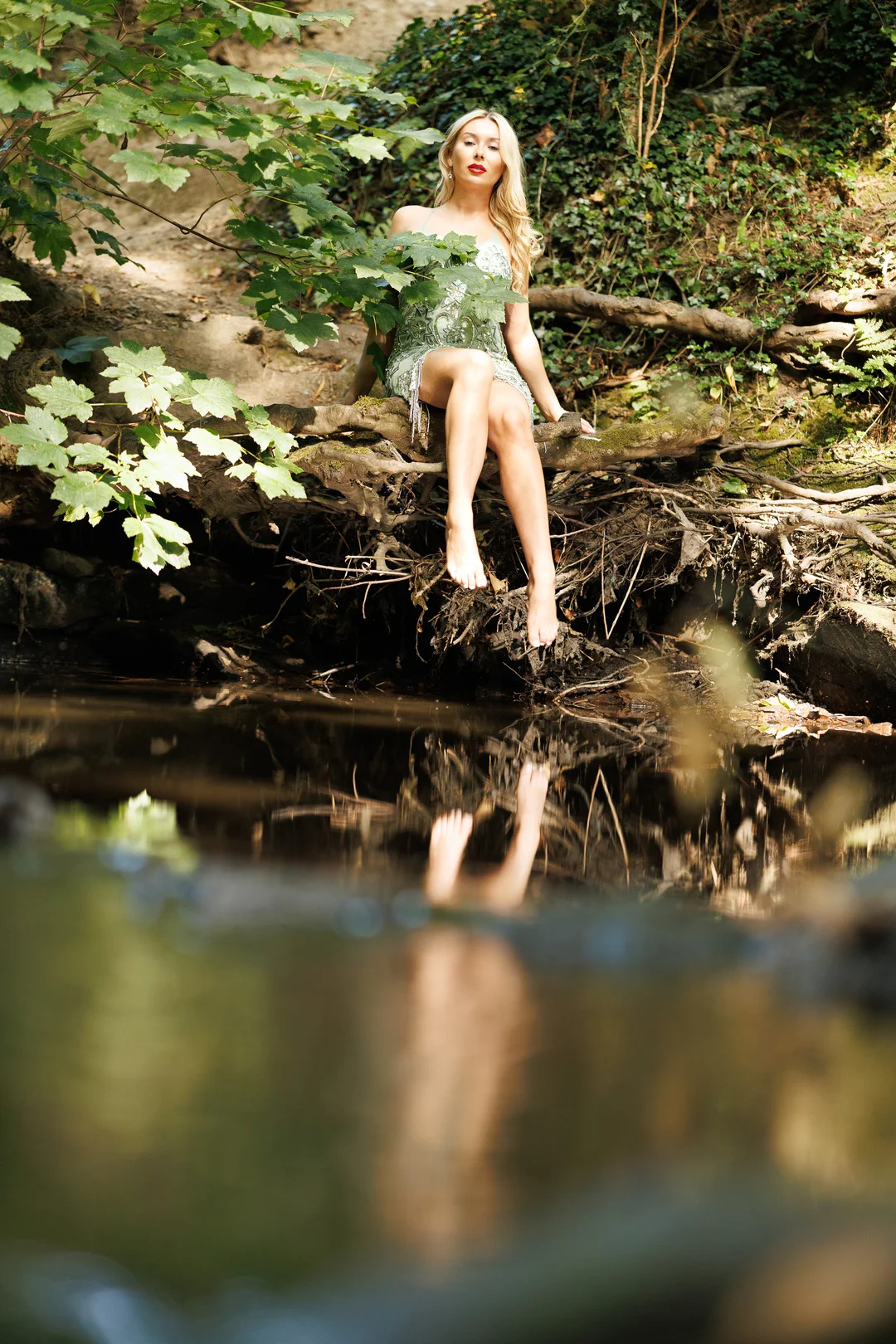 Blonde woman in a green dress sitting on a log by a stream with a reflection in the water.