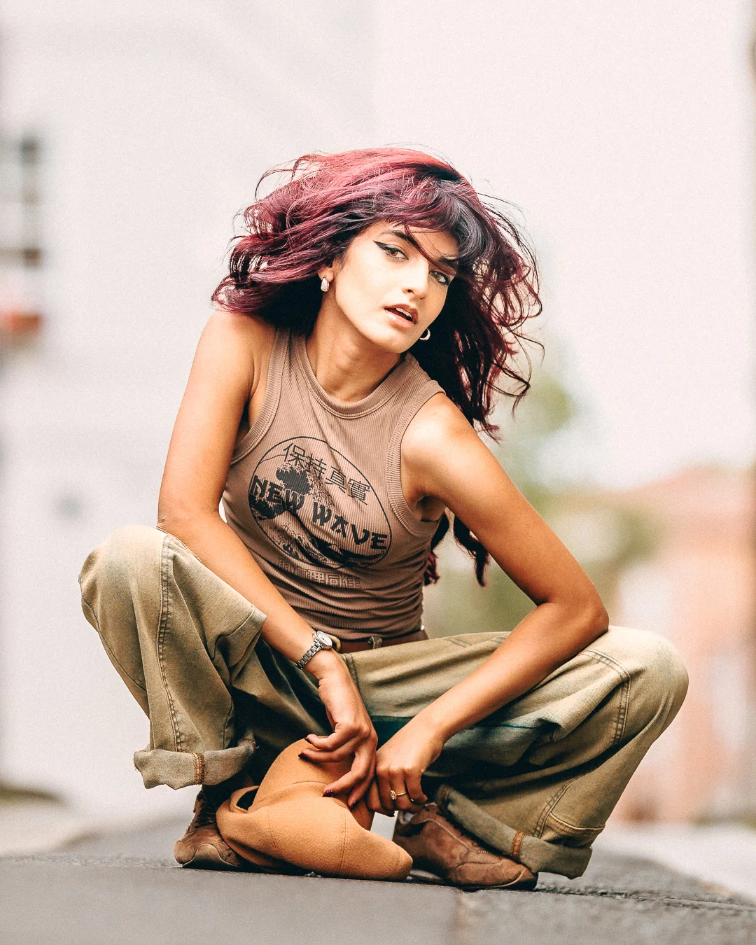 A woman with vibrant red hair in a tan crop top and khaki pants crouching on an urban street.