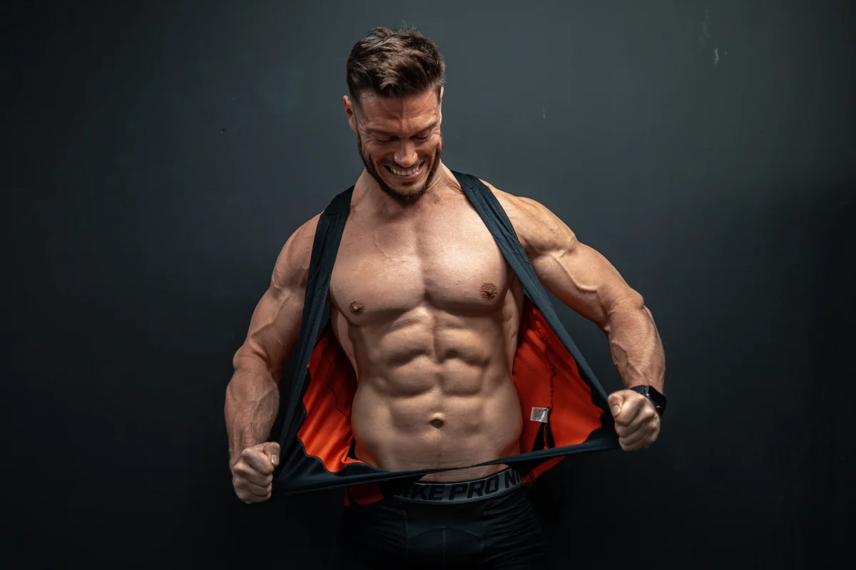A muscular man in a black and orange vest, smiling and pulling the vest open to reveal his abs against a dark background.