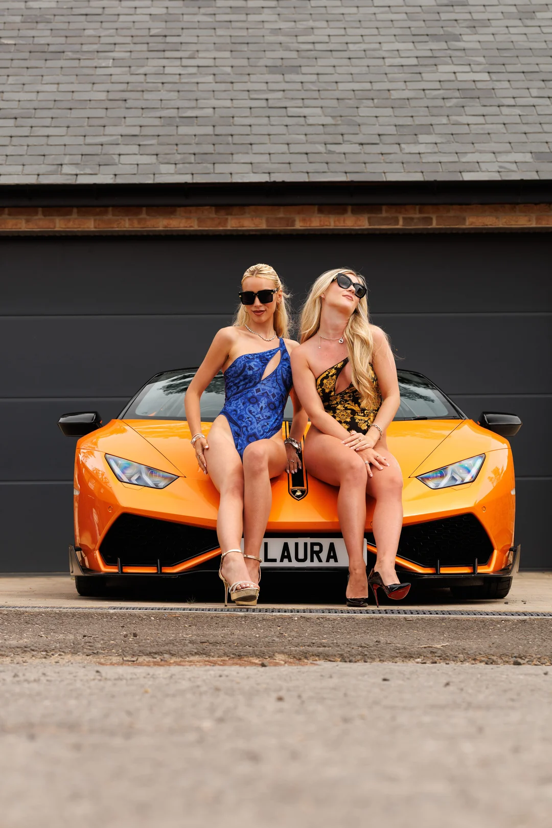 Two blonde women in revealing blue and gold swimwear posing on the hood of an orange Lamborghini.