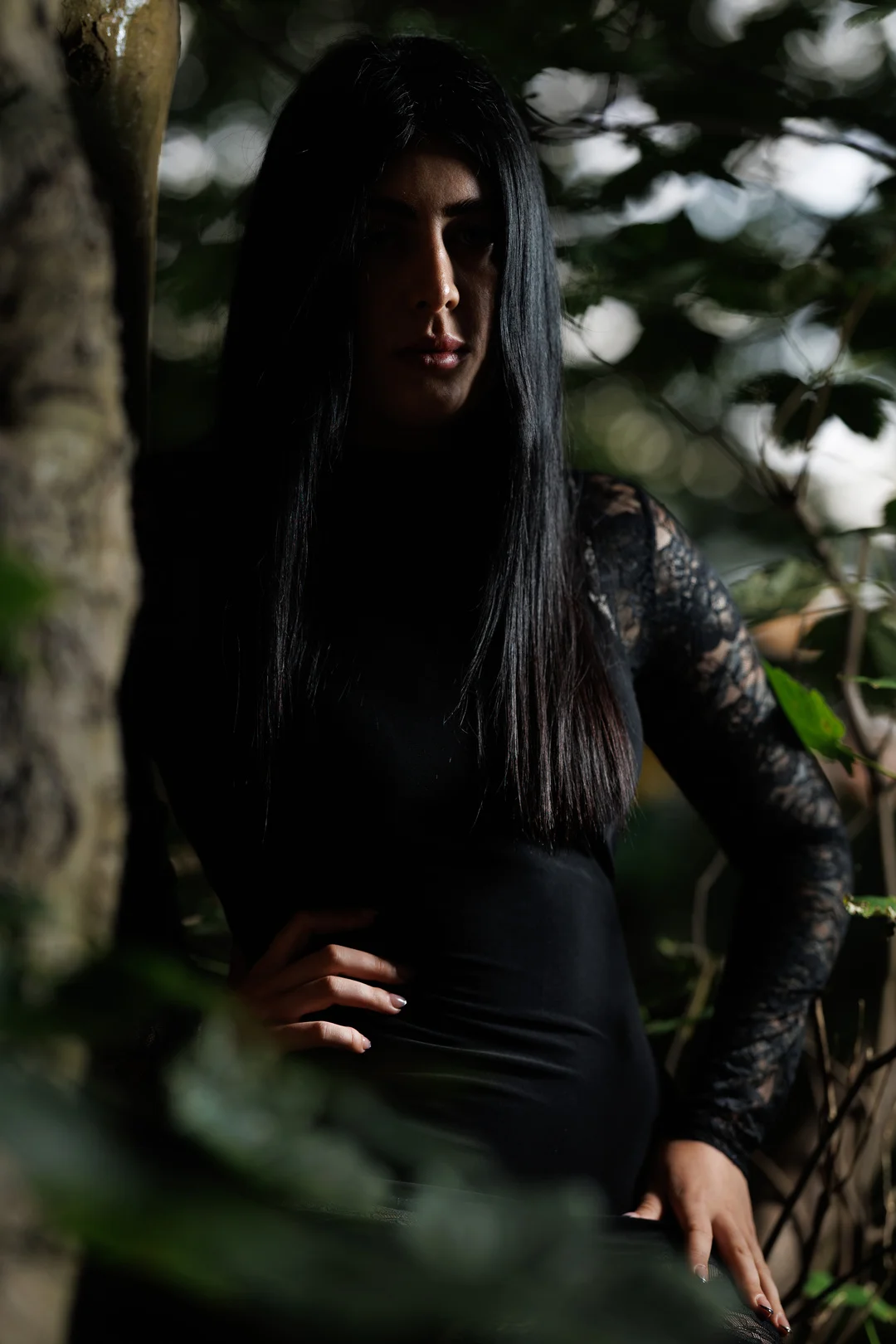 Moody portrait of a woman with long black hair in a black lace top, posing in a shaded forest.