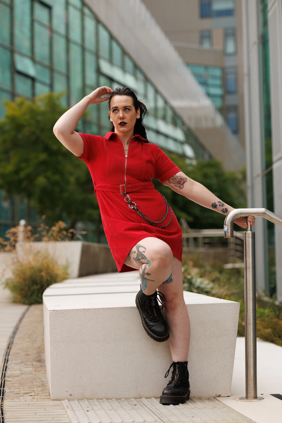 A person with tattoos in a red mini-dress and black boots posing outdoors against a modern glass building.