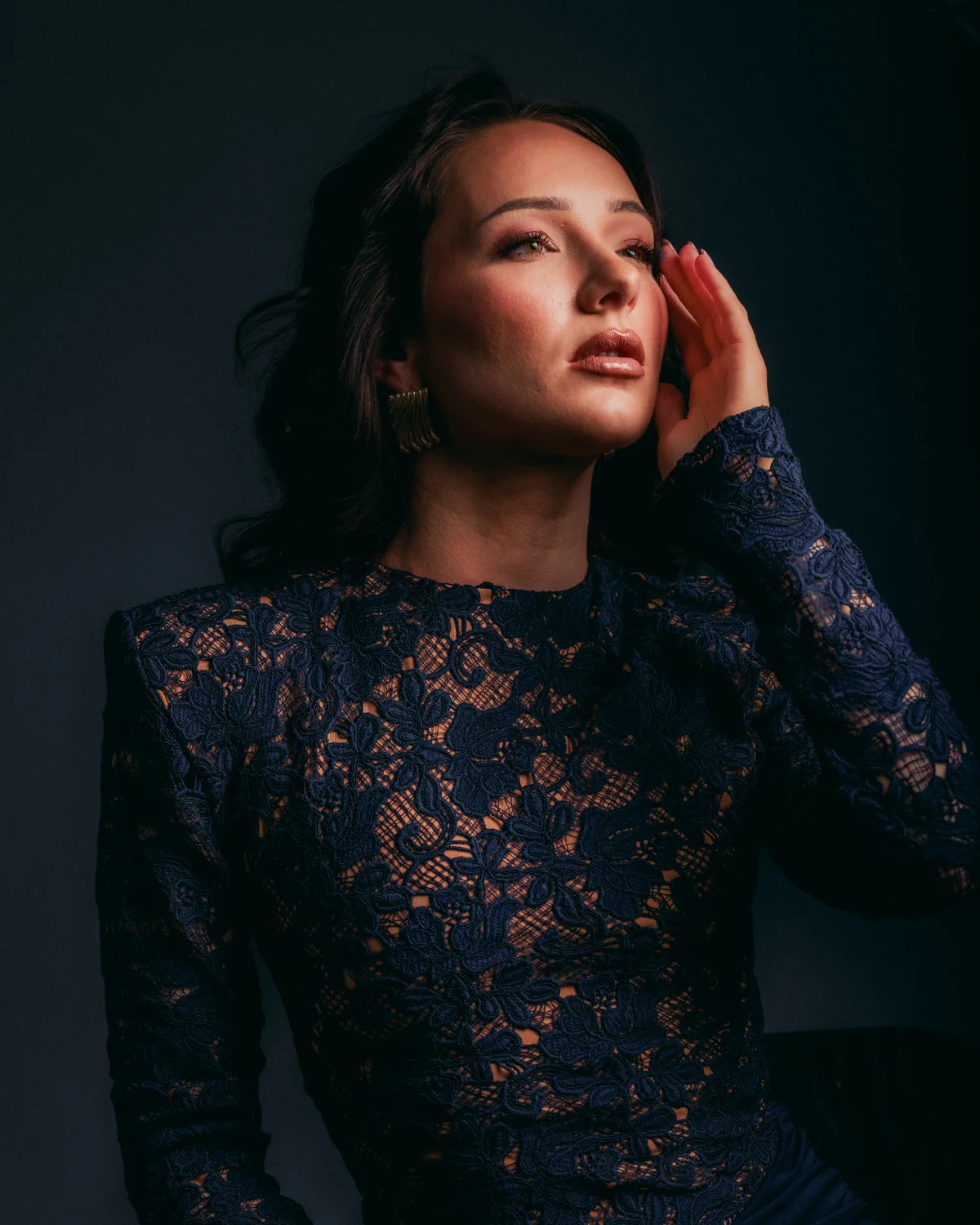Woman in a navy blue lace dress posing against a dark background with moody studio lighting.