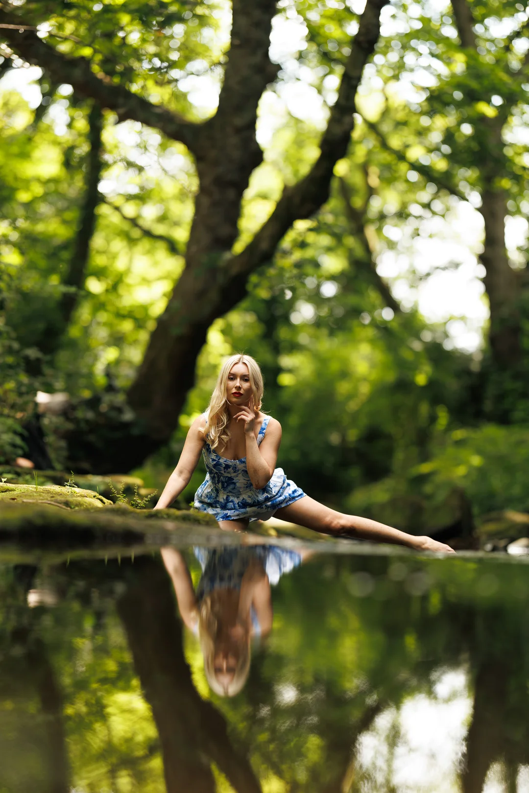 Blonde woman in a blue floral dress posing in a forest stream with a clear reflection.