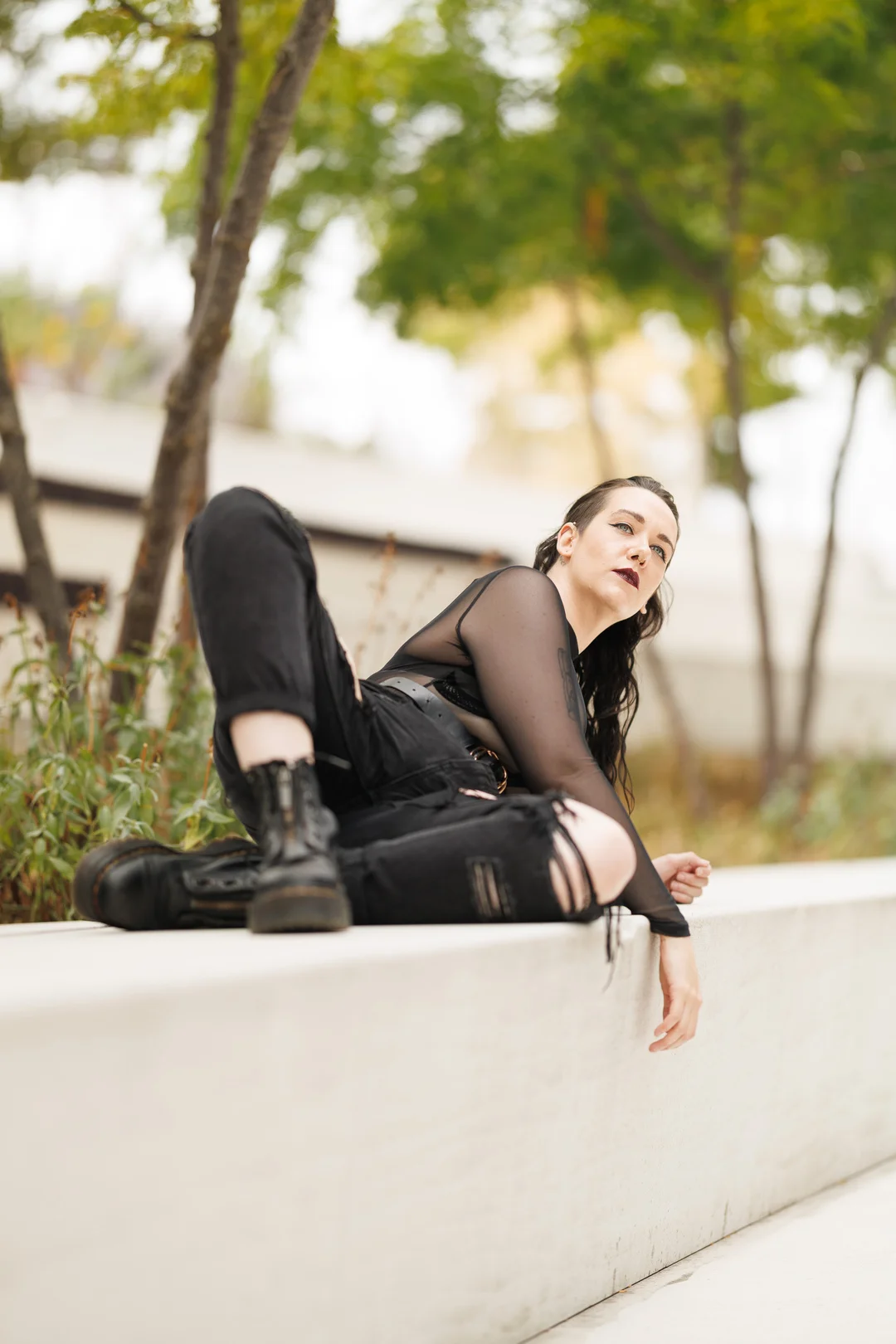 Person in edgy black sheer and ripped clothing reclining on a white concrete ledge outdoors.