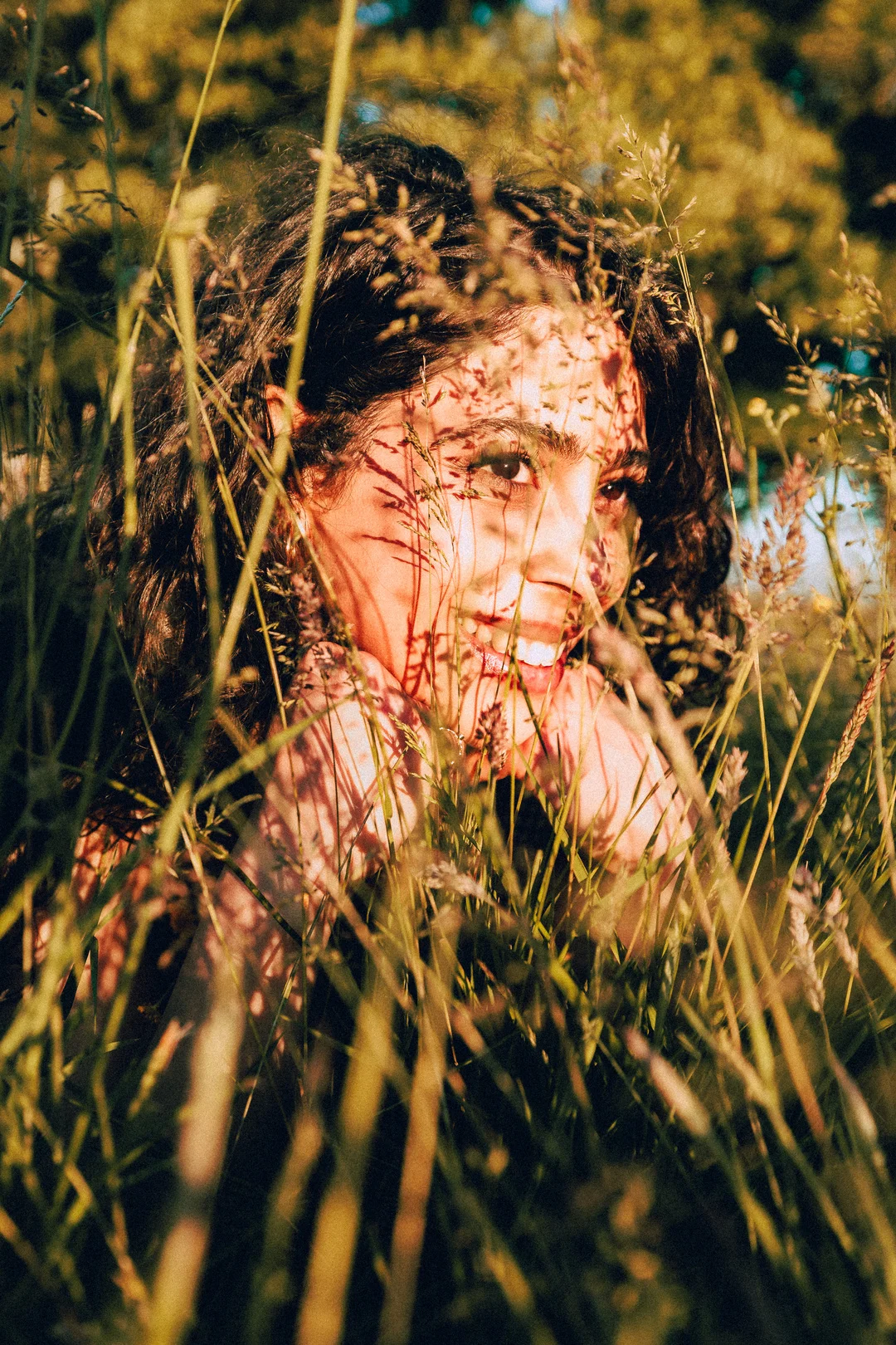 Close-up portrait of a woman partially hidden by tall grass with dappled sunlight on her face.
