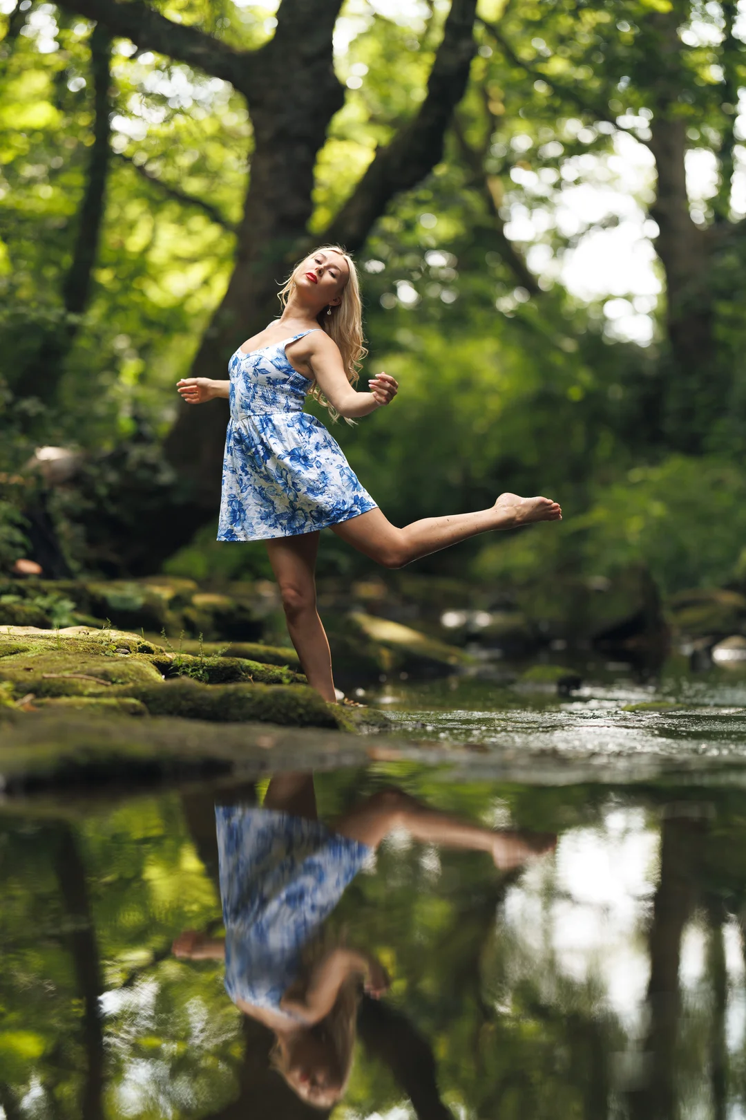 Woman in a blue floral dress posing on a mossy rock by a forest stream, with a reflection in the water.