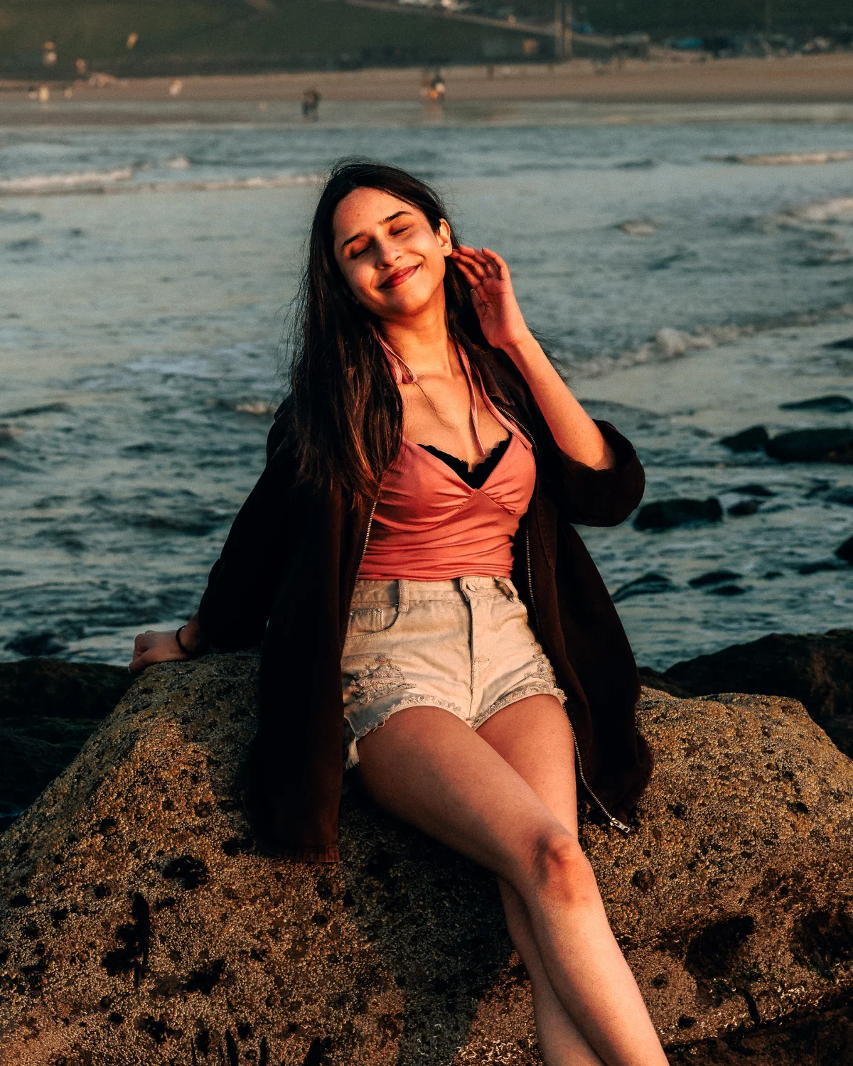 A woman with long dark hair sitting on a rock at a beach during golden hour, wearing a pink top and denim shorts.