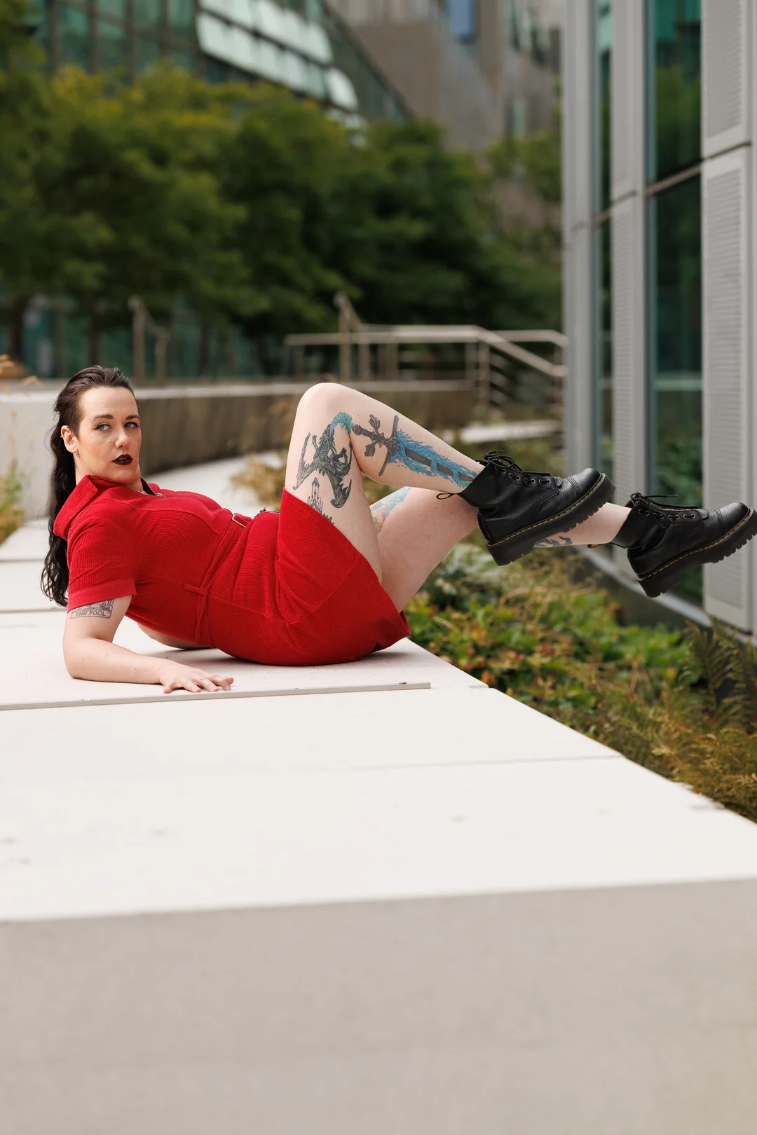 Woman in a red dress posing on a white concrete ledge outdoors with tattoos and black boots.