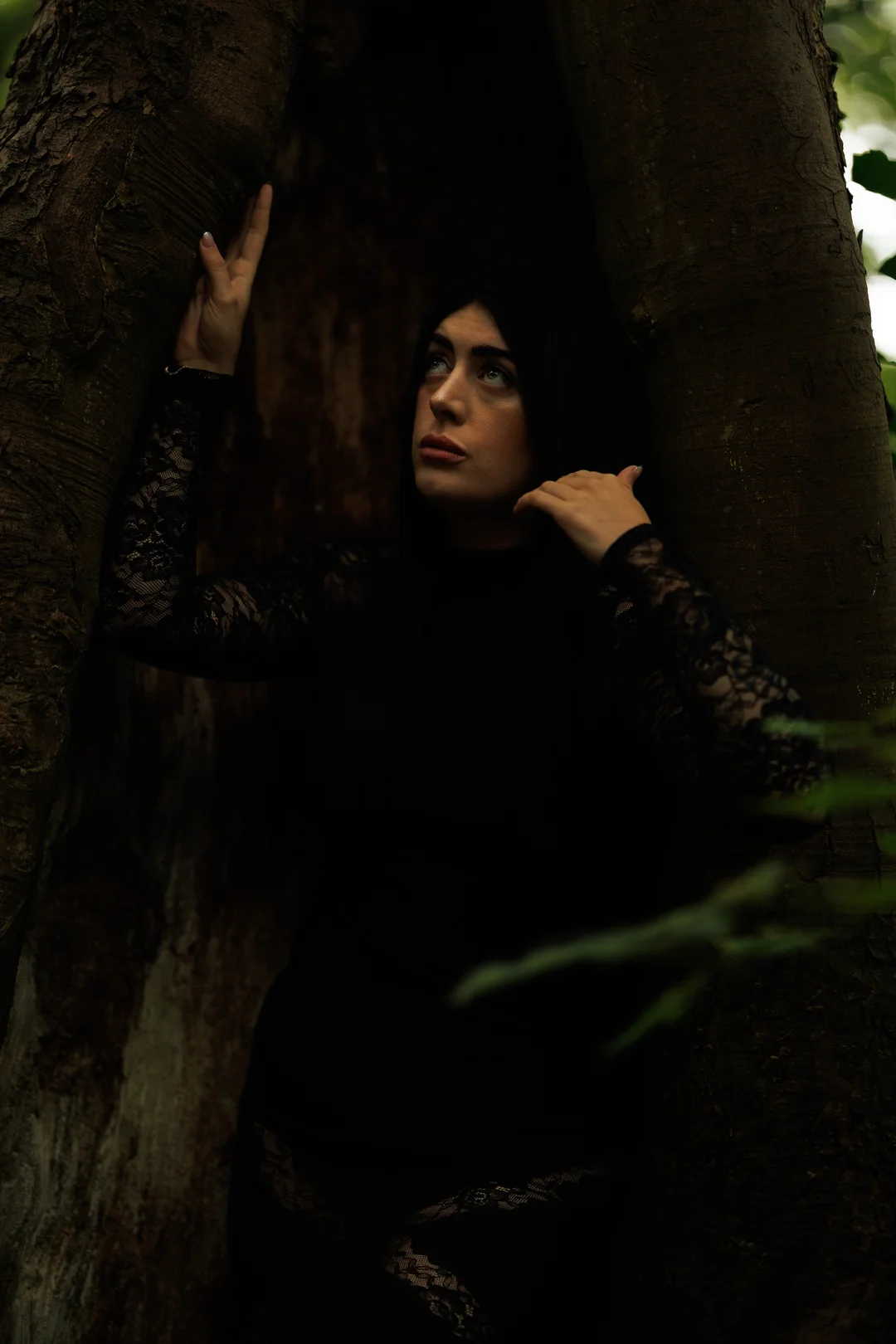 Woman in a black lace dress posing inside a hollow tree trunk, looking upwards with a moody expression.