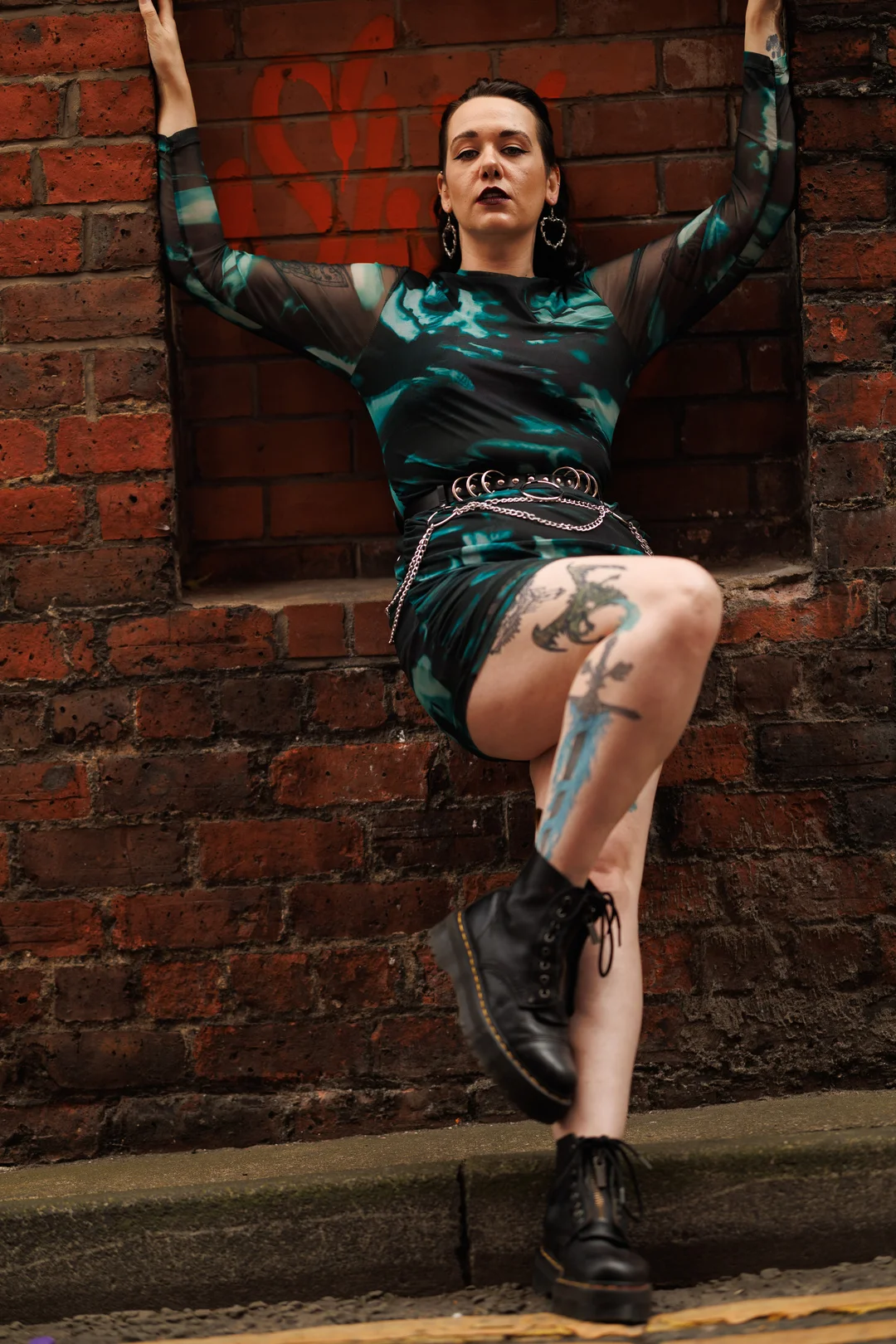 A person with tattoos in a teal and black outfit and black boots posing against a red brick wall.