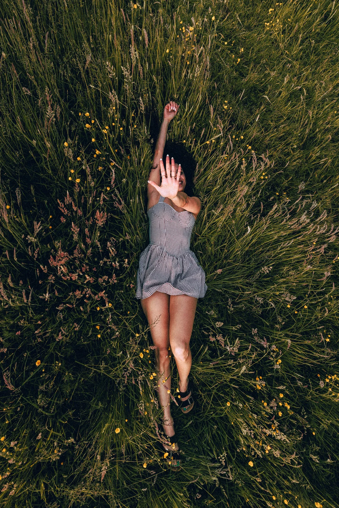 A woman in a gingham dress lying in a lush green field of grass and yellow wildflowers.
