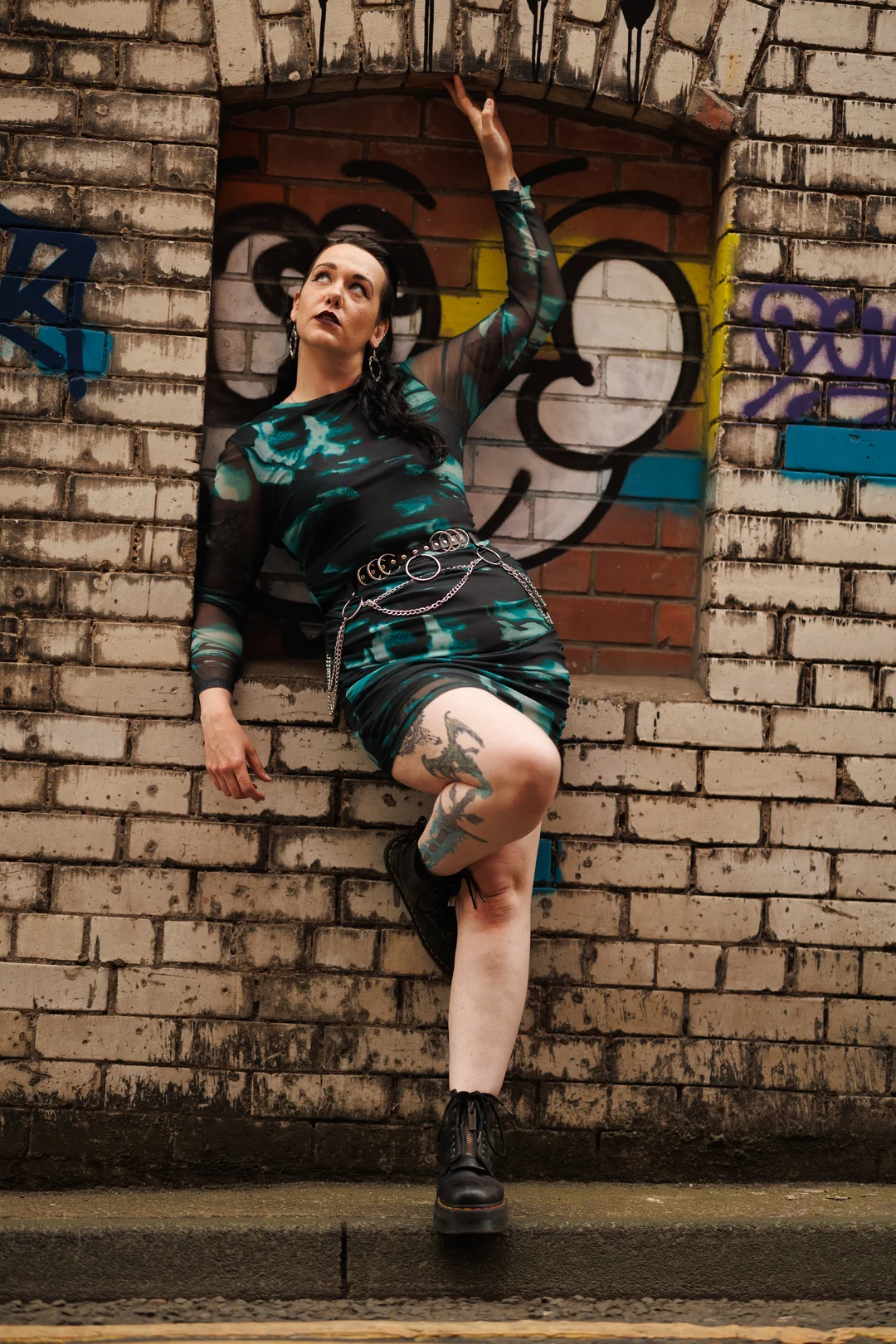 Woman in a teal and black patterned dress posing against a graffiti-covered brick wall.