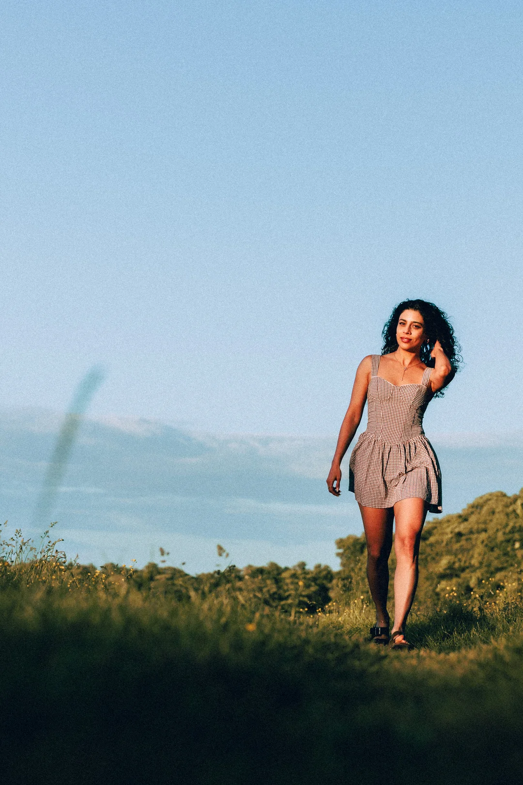 Woman with curly hair in a checkered mini dress walking through a grassy field under a clear blue sky.