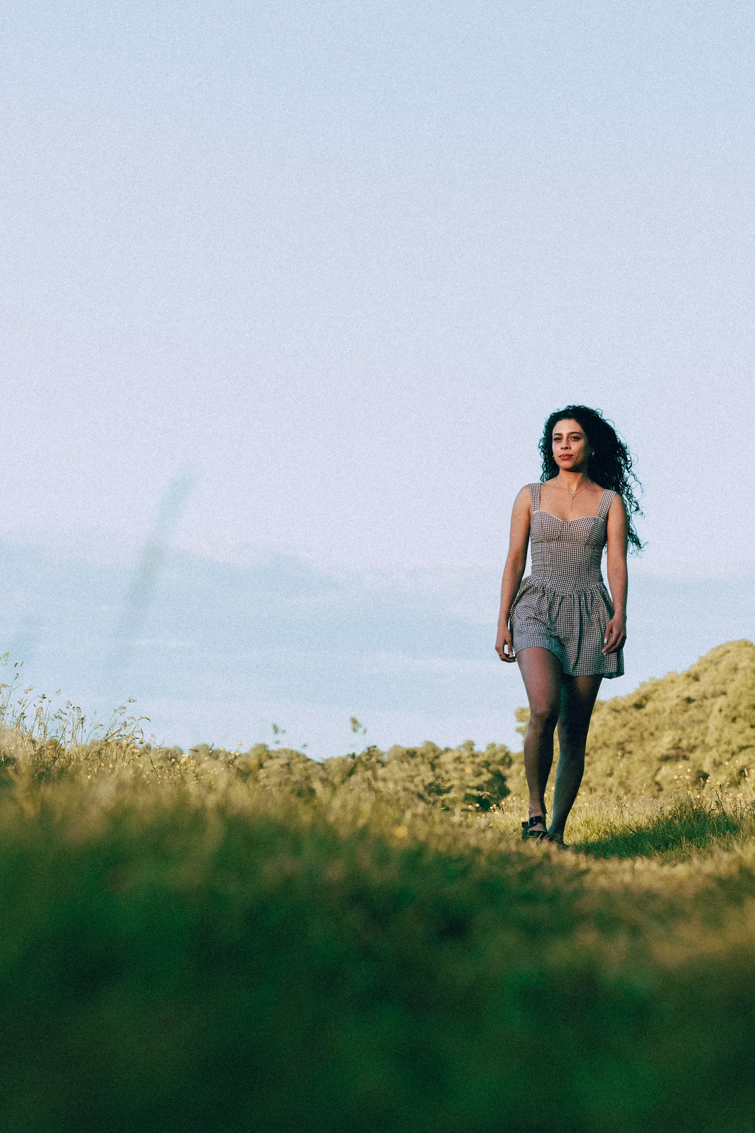 A woman with curly hair in a gingham dress standing in a grassy field under a pale sky.