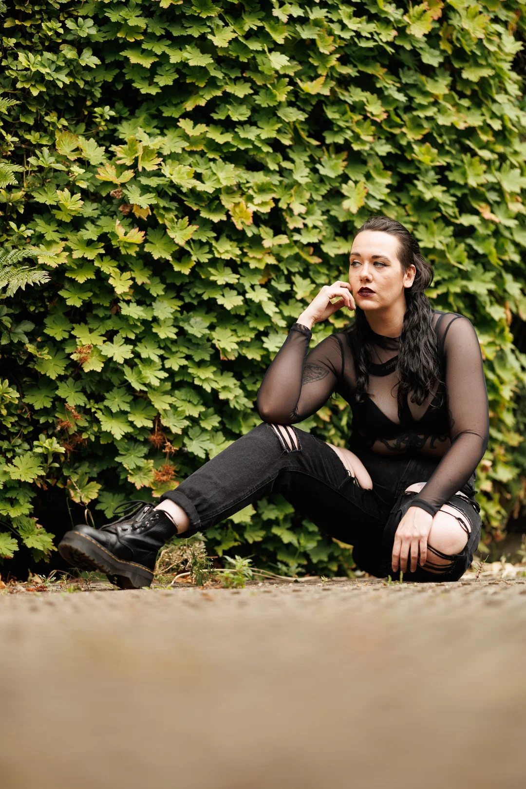Woman with dark makeup and hair sitting outdoors before a green hedge, wearing a sheer black top and ripped jeans.