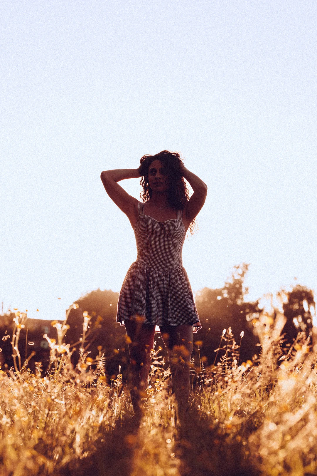A woman in a checkered dress posing in a golden field during sunset, hands behind her head.