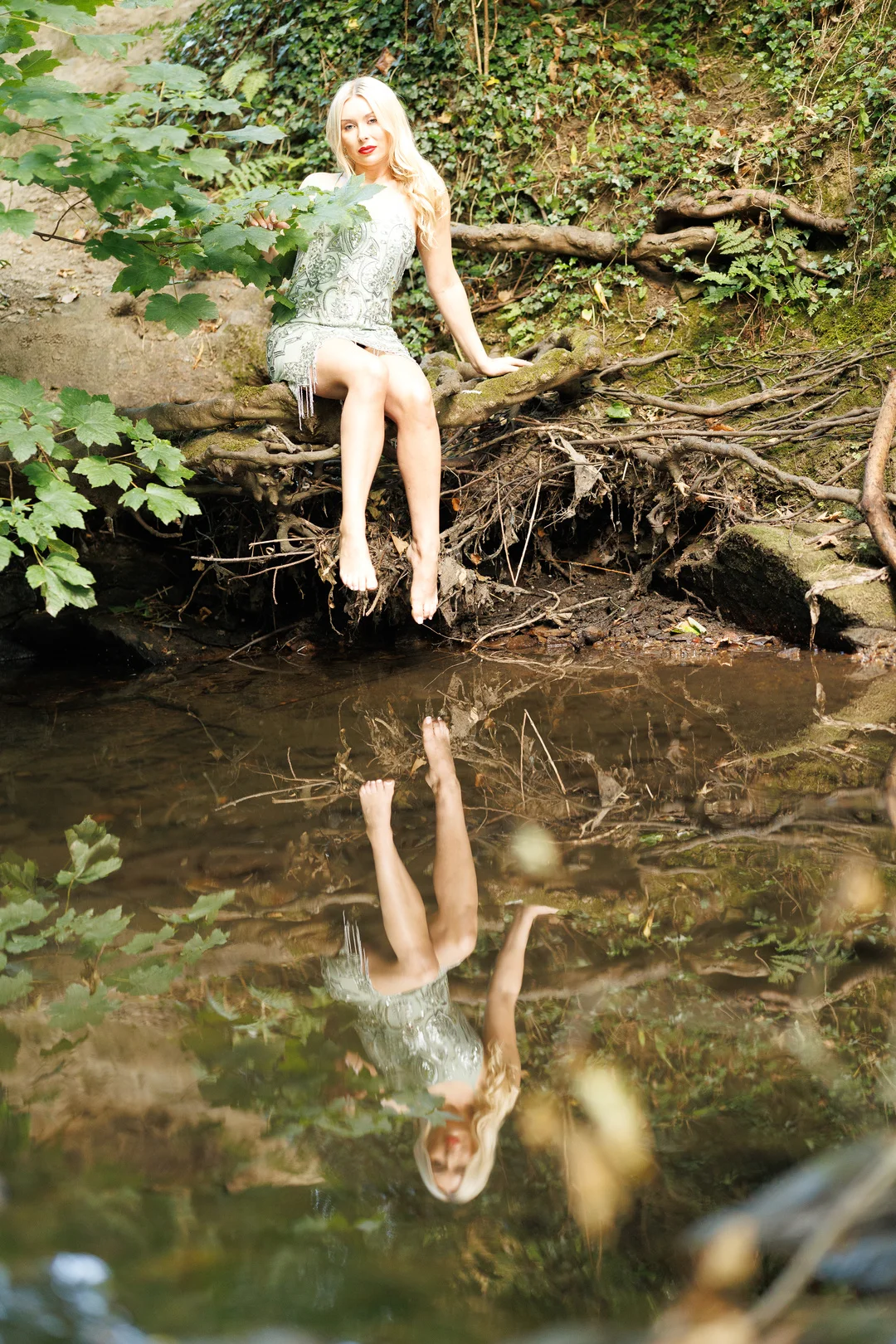 Blonde woman in a silver sequined dress sitting on a log by a stream, reflected in the water below.