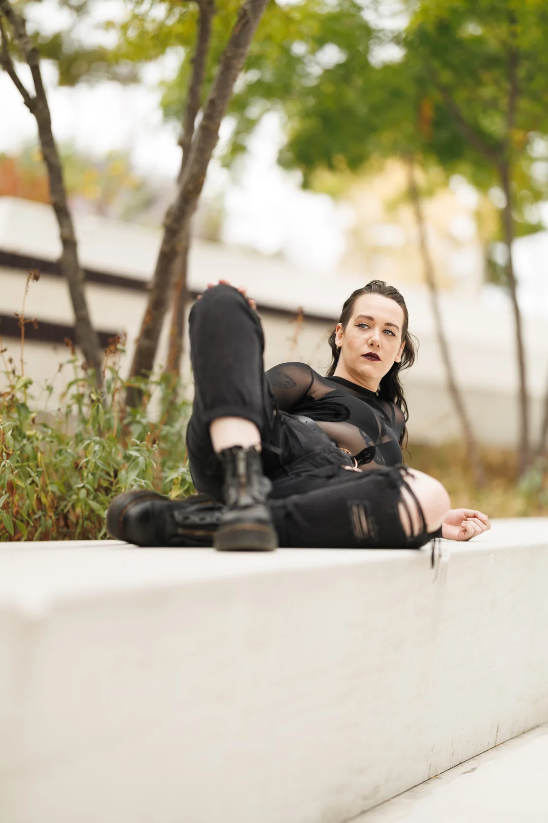 A person with dark makeup and black sheer clothing reclining on a white concrete ledge outdoors.