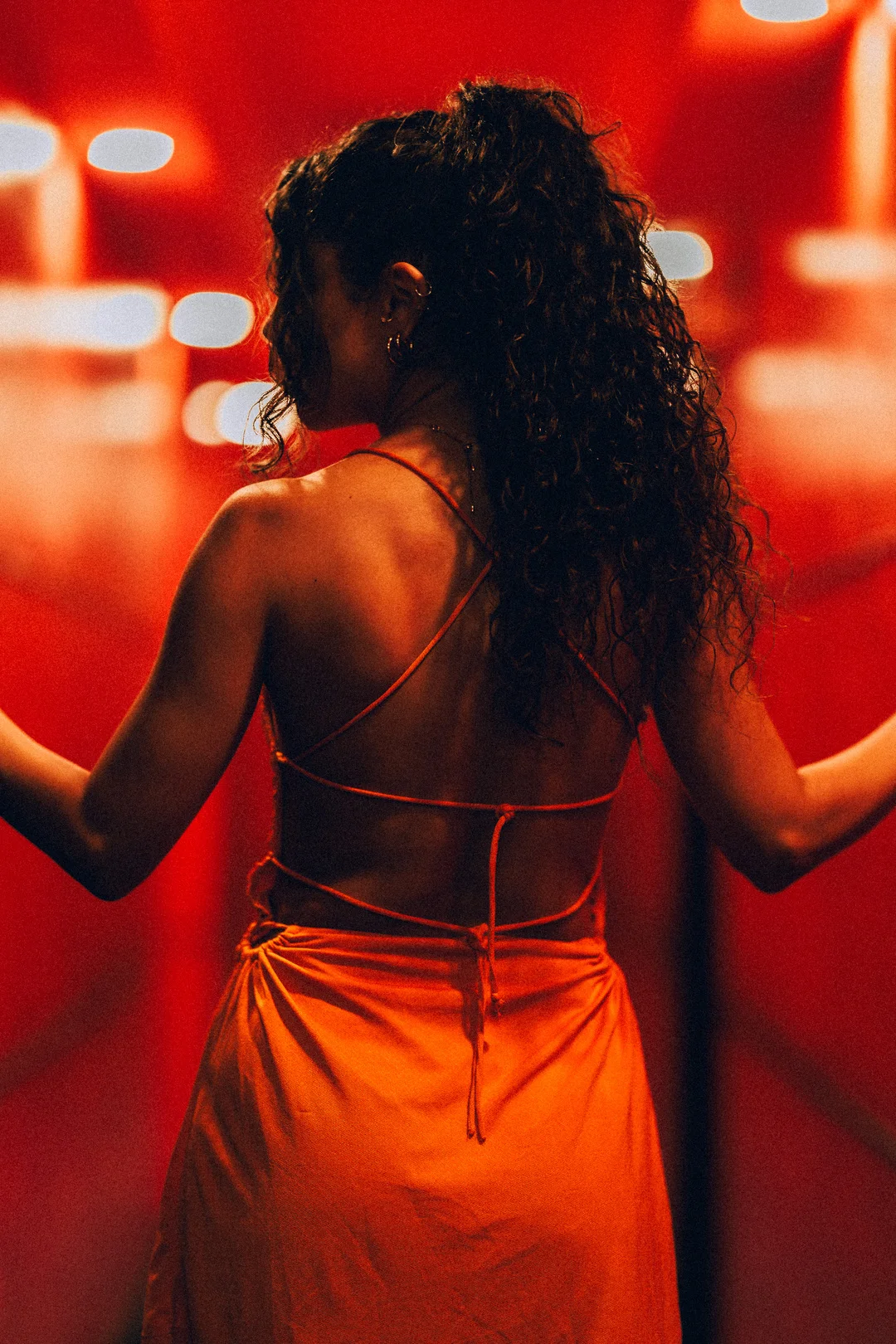 Woman with curly hair in an orange backless dress against a moody red background with bokeh lights.