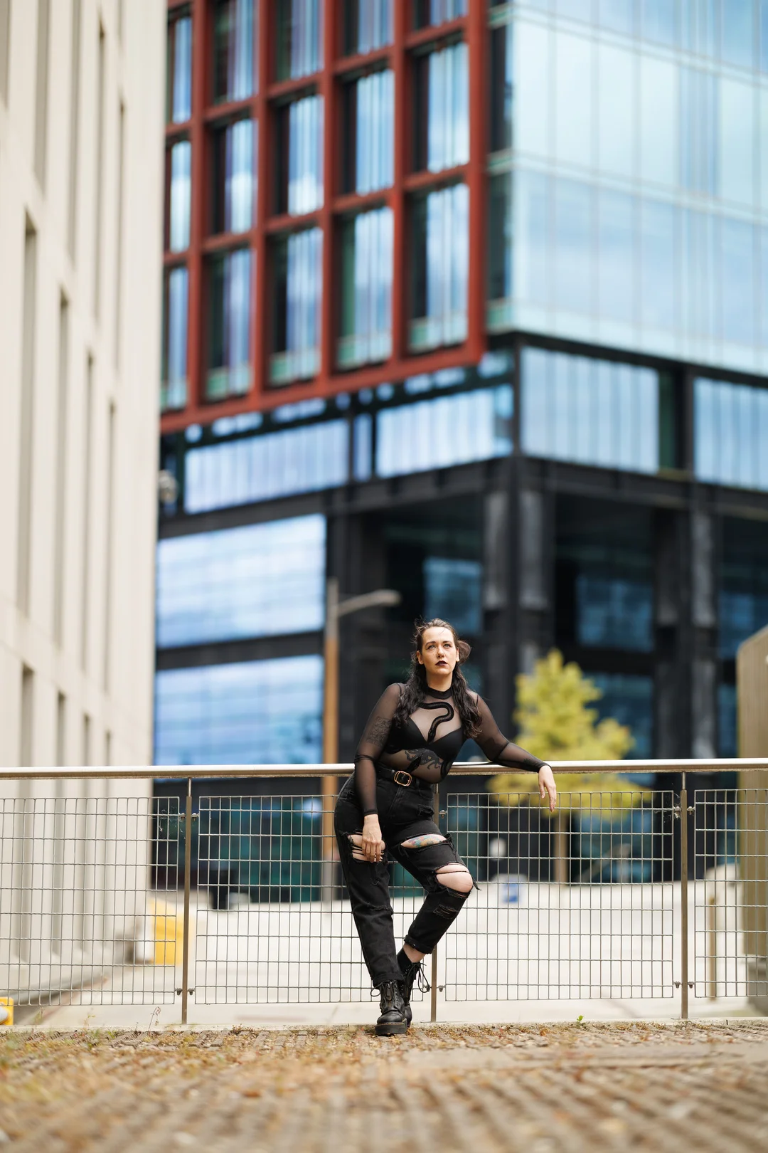 Woman in a sheer black top and ripped jeans leaning on a railing in an urban city setting.