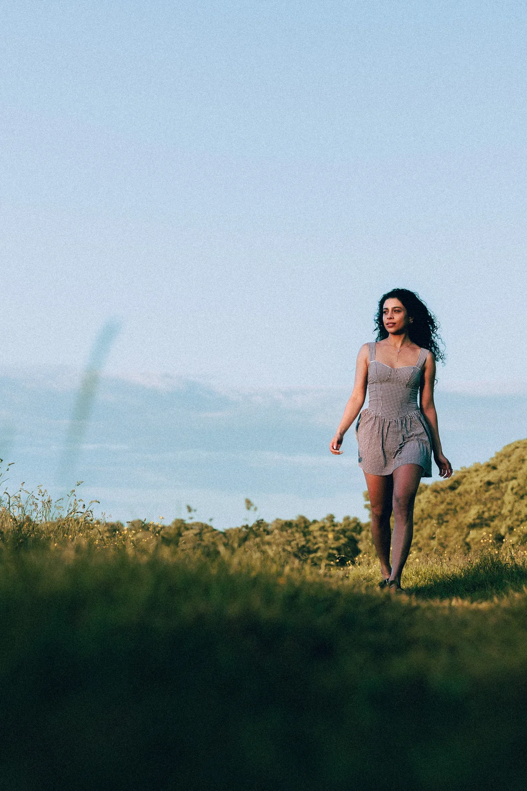 A woman with curly hair in a gingham dress walking through a grassy field under a clear blue sky.