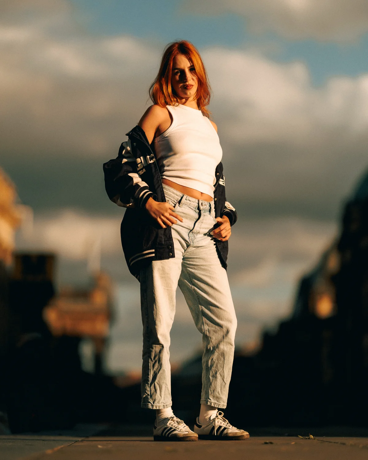 Young woman with red hair posing outdoors in a white crop top, light-wash jeans, and a navy varsity jacket.