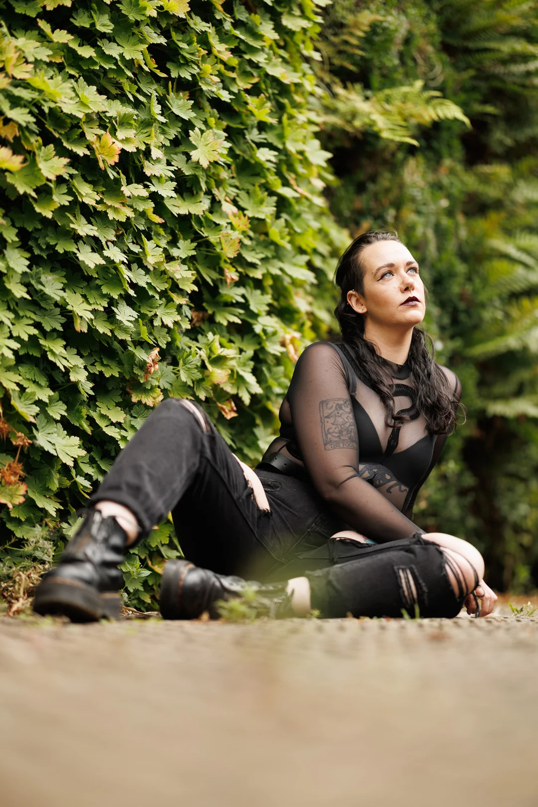 A person with dark hair and makeup, wearing a black mesh top and ripped black jeans, sitting before a green hedge.