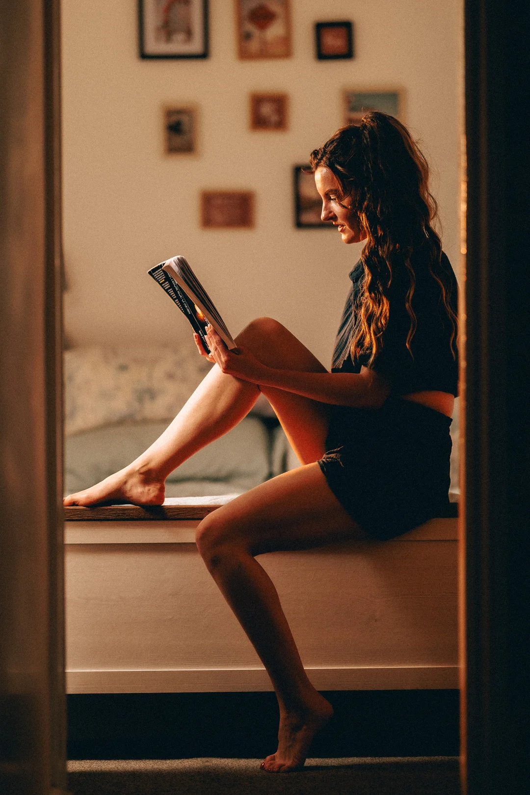 Woman sitting on a bed reading a book in warm, moody indoor lighting.