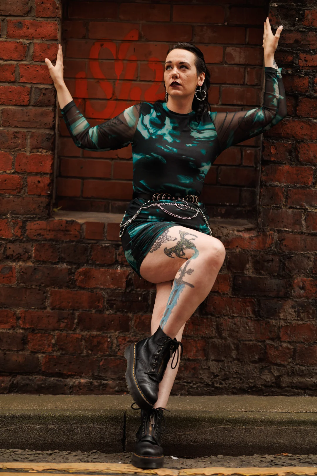 Person with tattoos in a teal and black dress and black boots posing against a red brick wall.