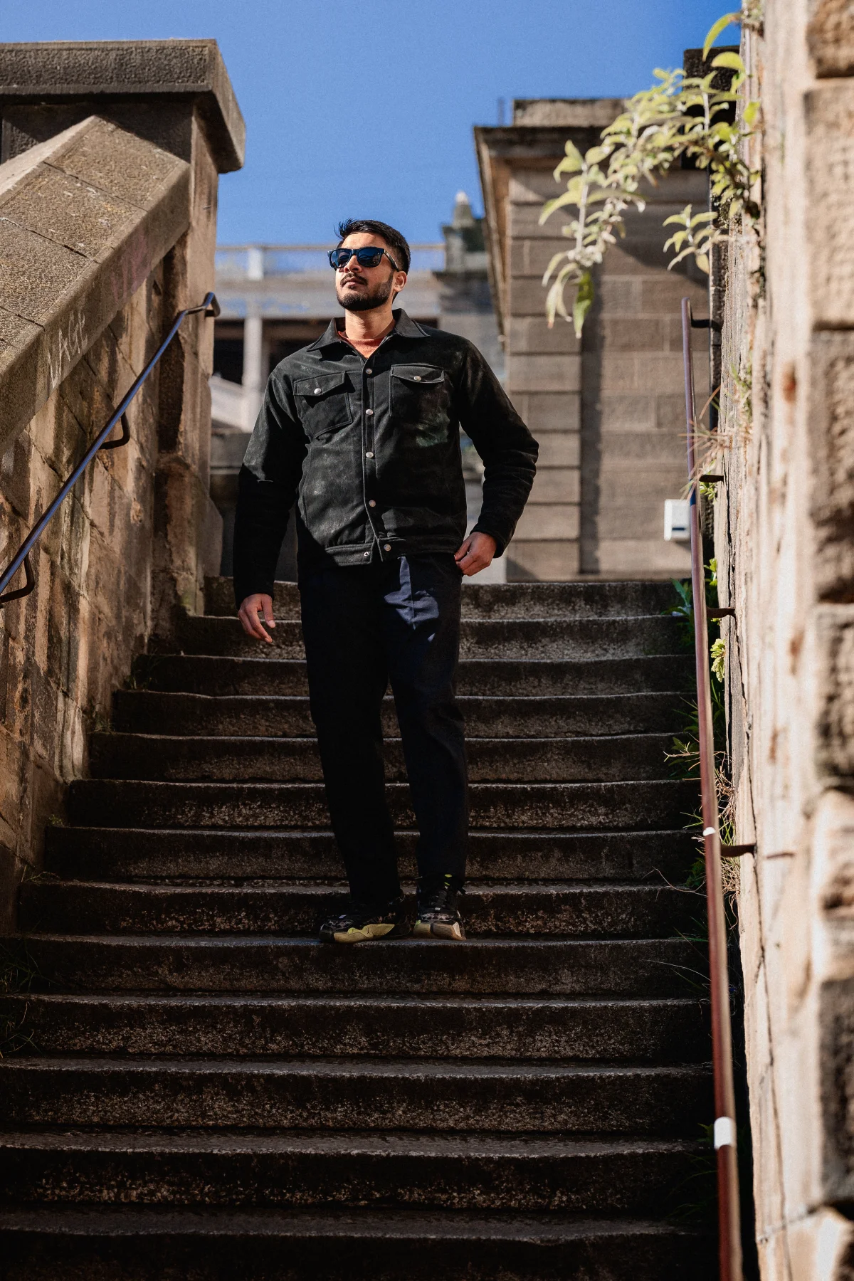 A man in a black jacket and sunglasses posing on stone stairs under a clear blue sky.